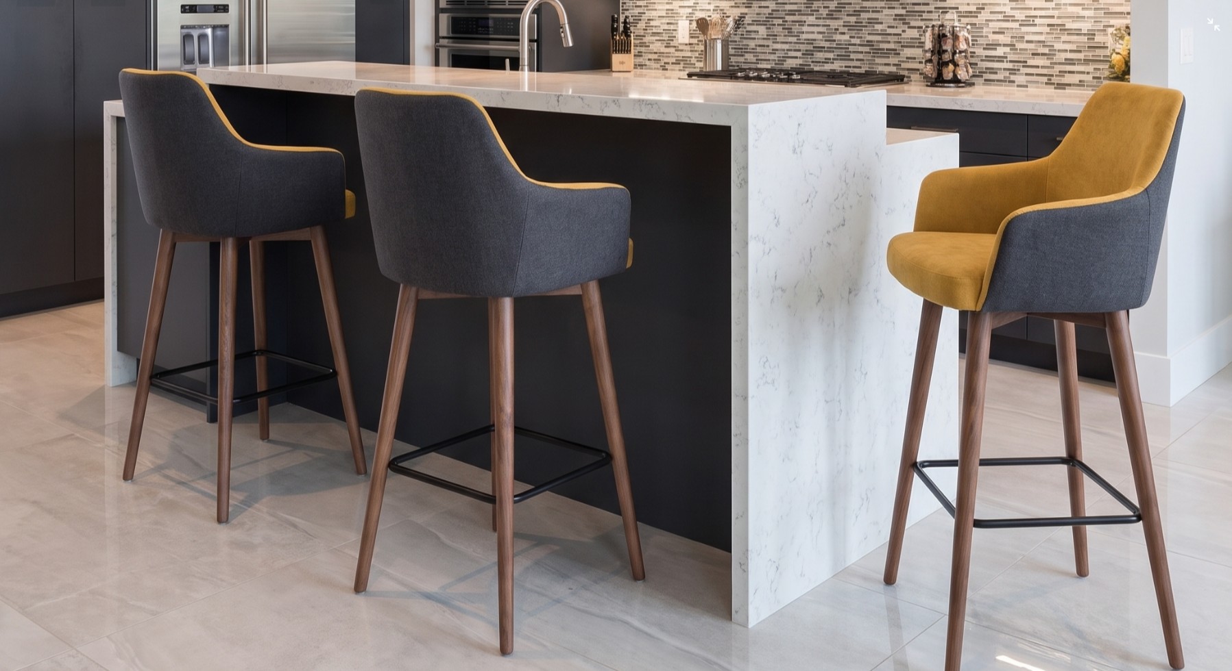 Taburetes altos con brazos tapizados en mostaza y gris, patas de madera y reposapiés negro junto a una isla